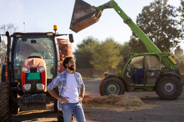 Çiftçi çiftlikte sığır beslerken traktör, mikser ve yükleyicinin önünde duruyor