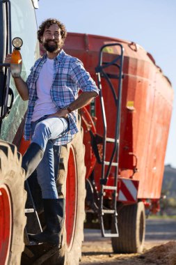 Çiftçi traktörün merdivenlerinde dururken gülümsüyor, arka planda yem karıştırıcı ile sığır çiftliğinde çalışmaya hazır.