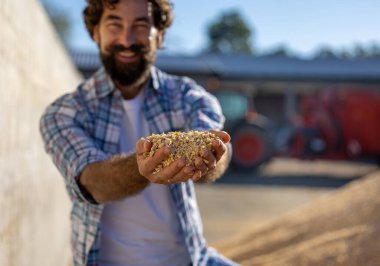 Erkek çiftçi, arka planda bir traktör ve besleyici mikserle bir çiftlikte bir kazığın yanında dururken elinde mısır tarlasını besliyor.