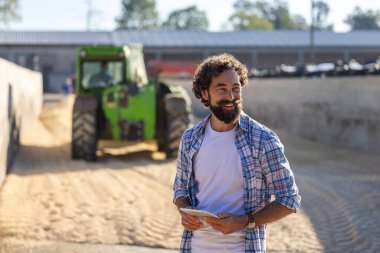 Traktörün önünde elinde tablet tutan çiftçi güneşli bir sonbahar gününde sığır çiftliğinde çalışıyor.