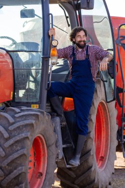 Tulumlu gülümseyen çiftçi traktörde rahatlıyor, çiftlik atmosferinin tadını çıkarıyor. Sahne, kırsal kesimde güneşli bir günü yakalıyor.