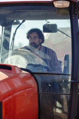 Sakallı ve bıyıklı çiftçi çiftlikte güneşli bir günde traktörü açık mavi gökyüzünün altına sürüyor.