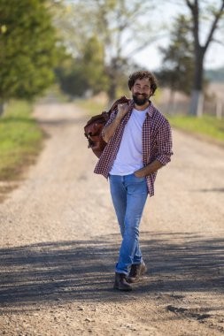 Gülümseyen adam, yeşil ağaçlar ve tarlalarla çevrili, omzunun üzerinde deri çanta taşıyan, kırsal alanda çakıl yolda yürüyor.