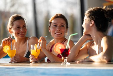 Üç kadın havuzda dinleniyor, gülüyor ve sıcak havanın tadını çıkarırken renkli kokteyller yudumluyorlar. Enerjik atmosfer onların neşeli ruhlarını güçlendirir.