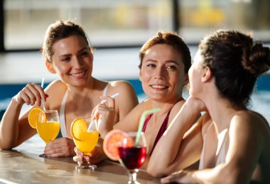 Üç kadın havuzda dinleniyor, gülüyor ve sıcak havanın tadını çıkarırken renkli kokteyller yudumluyorlar. Enerjik atmosfer onların neşeli ruhlarını güçlendirir.