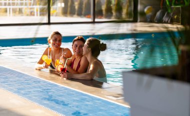 Üç kadın havuzda dinleniyor, gülüyor ve sıcak havanın tadını çıkarırken renkli kokteyller yudumluyorlar. Enerjik atmosfer onların neşeli ruhlarını güçlendirir.