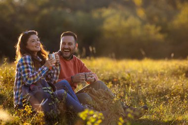 Sonbaharda güneşli çayırlarda iki insan birlikte rahatlar, sıcak içkilerini yudumlar ve gülümsemelerini paylaşırlar. Altın ışık onların neşeli anlarına sıcaklık katar..