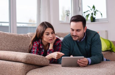 Genç çift kanepede oturuyor, tablette sörf yapıyor. Neşeli ifadeleri, sıcak oturma odasındaki ortak deneyimlerini yansıtıyor..