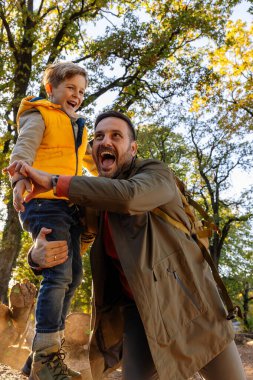Bir baba, gülümseyen oğlunu renkli sonbahar ağaçlarıyla çevrili güneşli bir parkta kaldırır. Mutluluk ve bağlantı anlarını paylaşıyorlar..