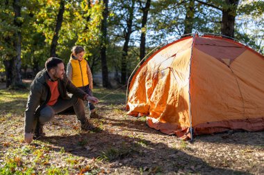 Bir baba ve oğlu, güneşli bir sonbahar gününde açık bir ormanda parlak turuncu bir çadır kurmak için birlikte çalışırlar. Yapraklar renk değiştiriyor