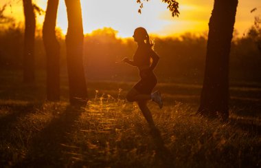 Gün batımında parkta koşan genç bir kadının silueti, ağaçlar ve sahneyi dolduran sıcak altın ışıkla çevrili.