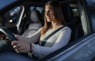 Genç bir kadın akşam boyunca araba sürerken gülümsüyor. Emniyet kemeri takıyor ve önündeki yola odaklanmış gibi görünüyor..