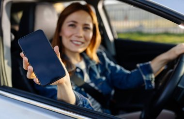 Bir kadın park halindeki bir arabada oturuyor ve akıllı telefonunu gülümseyerek kameraya doğru tutuyor. Gündüz oldu ve o mutlu görünüyor..