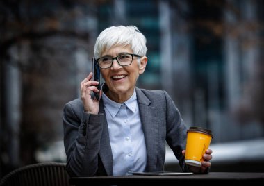 İş kadını açık bir masada oturup elinde kahve fincanıyla telefonla konuşuyor. Takım elbise giymiş ve kısa saçlı..