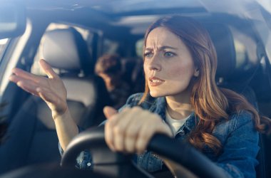 Hayal kırıklığını ifade ederken araba kullanan bir kadın. Arka koltukta bir çocuk görülebilir. Sahne gündüzleri çekiliyor..