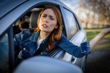 Bir kadın arabasının camından dışarı doğru eğilip konuşuyor. O, gün içinde bir parkta araba sürerken eliyle jest yapar. Arabanın içinde bir çocuk görülebilir..