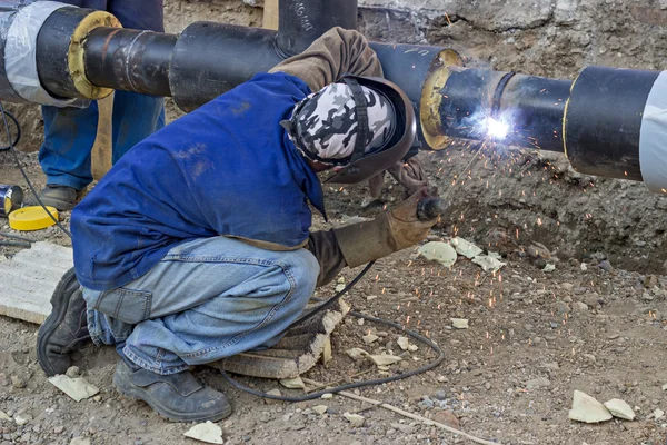 Welder welding underground steel pipe kneels on ground — Stock Photo, Image