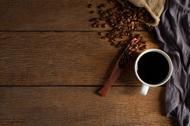 Beyaz seramik fincanda, siyah sıcak kahvenin üstünde kahve çekirdekleri ahşap masa arkasında çuval içinde kavrulmuş. Kopyalama alanı olan düz.