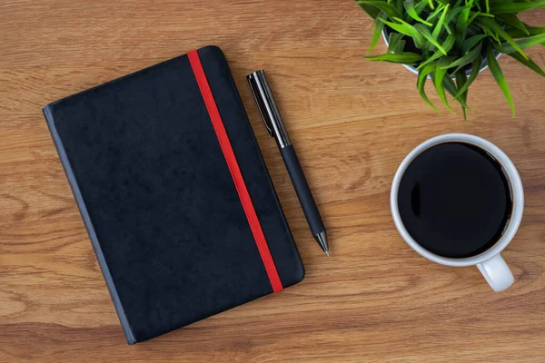 Top view of black closed cover blank notebook with coffee cup and pen for taking write notes on wood table background. Flat lay, creative workspace office. Business-education concept with copy space.