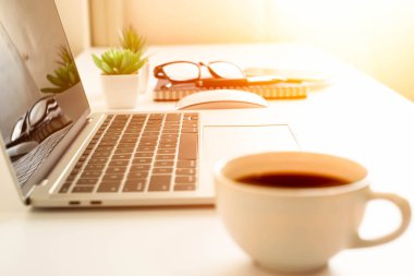 Masa başındaki dizüstü bilgisayarın yumuşak odağı, kahve fincanı, not defteri, bardaklar. İş ve finans kavramı. Penceresinde fotokopi alanı olan güneş ışığı olan işyeri.