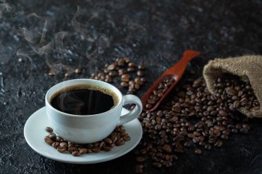 Sabah içeceği menüsü için dumanlı sıcak sade kahve, beyaz seramik bardakta kahve çekirdekleri koyu grunge kırsal masa arka planında çuval içinde kavrulmuş. Kopyalama alanı olan düz.