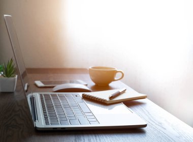Laptop bilgisayarı, kahve fincanı, not defteri ve bardaklarla dolu ofis masasının yumuşak odağı. İş ve finans kavramı. Penceresinden fotokopi alanına sahip sabah güneş ışığına sahip işyeri.