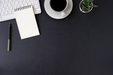 Siyah deri ofis masası. Bilgisayar beyaz klavyesi ve boş not defteri olan çalışma alanı diğer ofis malzemeleriyle dolu bir kalem. Kopyalama alanı olan üst görünüm, düz yatay.