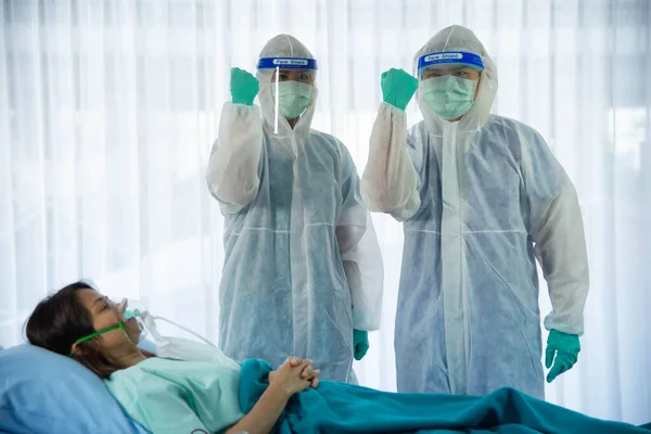 Two medical staffs with PPE suit and facial mask standing and hand up for fighting beside aging woman patient infected with CORONA VISRUS or COVID-19 bedroom in quarantine room at the hospital.