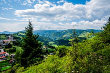 Yazın Asıago Platosu 'nun panoramik manzarası, yemyeşil tepeler, küçük dağlık köyler, ve bulutlu bir gökyüzü altında dramatik sıradağlar..