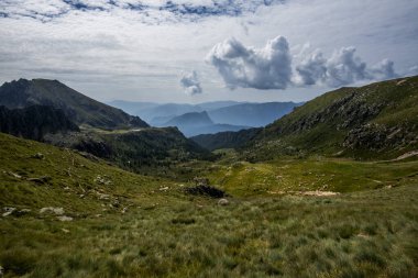 İtalya, Pieve Tesino yakınlarındaki Lagorai dağları, katmanlı mavi tepeler, dramatik bulutlar ve uzak otlayan sürülerle benekli yeşil alp yamaçları..