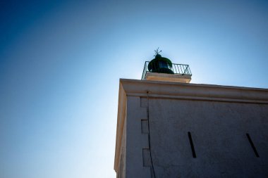 Korsika 'da yeşil tepeli bir deniz fenerinin yakın görüntüsü, açık mavi gökyüzüne karşı çekilmiş, geometrik çizgiler ve parlak yaz ışığı..