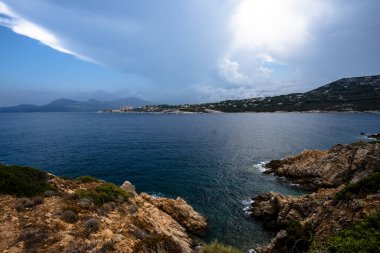 Fransa, Korsika 'da dramatik bulutların altında Akdeniz' in panoramik manzarası ve uzak kıyı köyü. Vahşi kıyı şeridi, derin mavi su ve atmosferik ışık.