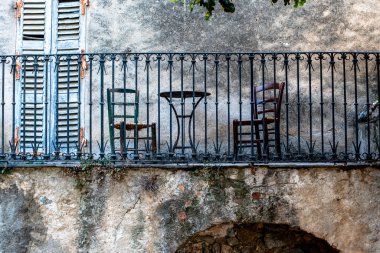 Geleneksel Akdeniz balkonunda demir parmaklıklar, tahta sandalyeler ve korsika, Fransa 'da eskimiş panjurlar. Dengeli kompozisyon, yumuşak renkler ve zengin dokular otantik köy mimarisini ön plana çıkarıyor.