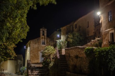 Taştan evler, kilise cephesi ve sokak lambalarıyla aydınlatılmış merdivenleri olan tarihi bir Korsikalı köy sokağı gece görüşü. Sessiz Akdeniz atmosferi, Avrupa mirası seyahat sahnesi.