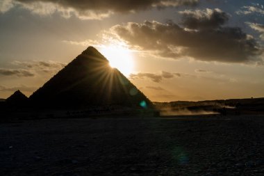 Kahire, Mısır 'da günbatımında eski bir piramidin silueti, güneş ışınları, sürüklenen toz ve deve binicileri ile dramatik bir tarih, ölçek ve çöl atmosferi yaratıyor..