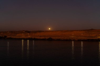 Aswan 'daki Nil Nehri' nin gece manzarası. Ay çöl tepelerinin üzerinde asılı duruyor. Suyun üzerine hafifçe yansıyor ve manzaranın sakinliğini vurguluyor..