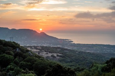 Güneşin doğuşuyla birlikte tepeler, bitki örtüsü ve bulutlu bir gökyüzünün altında sakin bir deniz manzarası. Seyahat, turizm, doğa ve hedef kullanımına uygundur.