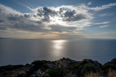 Kayalık bir sahil kayalığından, katmanlı bulutların ve geniş gökyüzünün altındaki sakin açık denizi yansıtan güneş ışığını görmek. Seyahat, doğa ve kıyı çevre kavramları için uygundur..