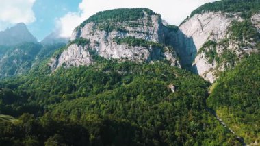 İsviçre Alpleri 'nin sarp kayalıkları yemyeşil ormanlarla kaplı güzel manzarası. Alçak bulutlar açık mavi bir gökyüzünün altında dağ zirveleri etrafında sürüklenirler. Alp yabanda bir yaz günü, İsviçre doğasının saf güzelliğini ve gücünü sergiliyor..