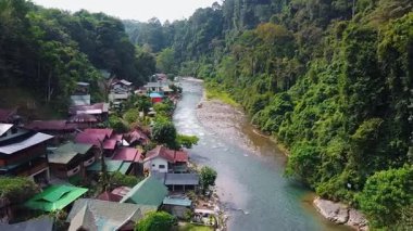 Bukit Lawang, Sumatra 'daki Bahorok Nehri' nin üzerinden geçen insansız hava aracı görüntüleri. Geleneksel köy evleri sağ taraftaki nehir kıyısında sıralanırken, yoğun tropikal yağmur ormanı sol taraf boyunca uzanır..