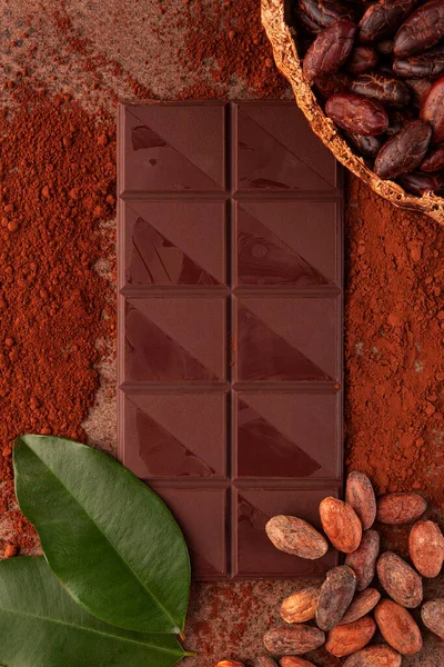 Cocoa beans in pod, theobroma cacao leafes and  chocolate bar flat lay on table. Delicious dark chocolate background.