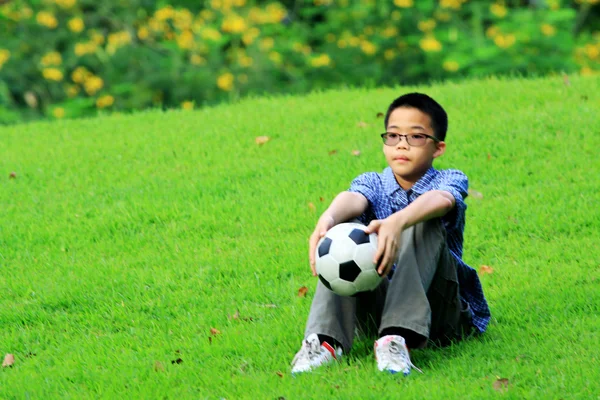 Asiático menino sentar na grama e segurar o futebol na mão — Fotografia de Stock