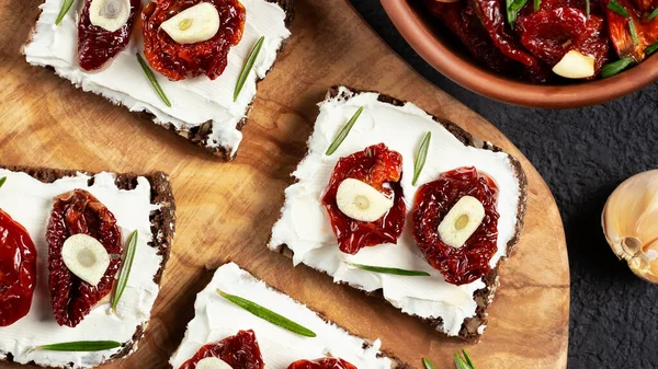 Sandwiches caseros de pan multigrano con queso crema y tomates secados al sol en una bandeja de madera. Concepto de alimentación saludable. Plano, vista superior — Foto de Stock