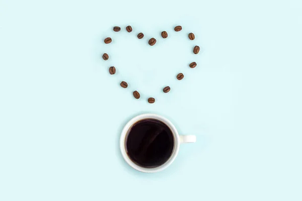 Creative composition: good morning or I love coffee. White cup with black coffee espresso and heart made from beans on blue background. Caffeine boost drink.