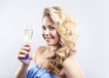 Bir bardak şampanyayla güzel kıvırcık saçlı sarışın. Mavi gece elbisesi giymiş genç bir kadın. Gri bir arka plan üzerinde gülümseyen kız