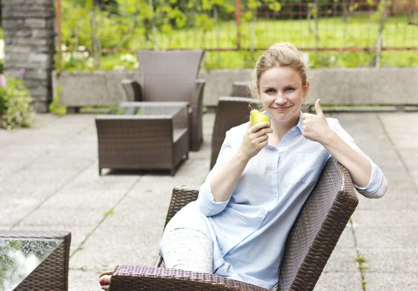 Hermosa rubia comiendo una pera. Jovencita sonriente sentada en la terraza. Hermosa mujer con camisa azul. Una joven lleva un estilo de vida saludable — Foto de Stock