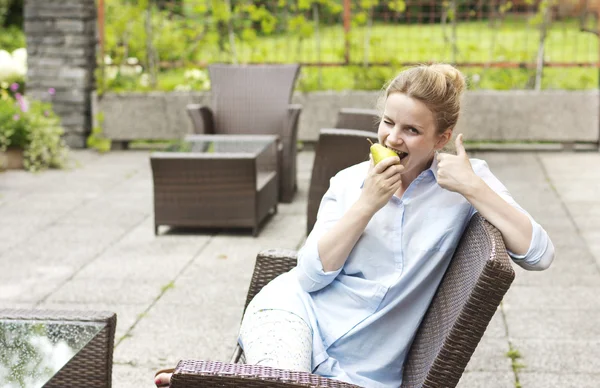 Mooie blonde eten van een peer. Lachende meisje zittend op het terras. Mooie vrouw in een blauw shirt. Een jong meisje leiden een gezonde levensstijl — Stockfoto