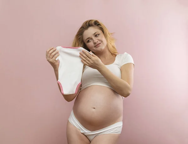 Giovane Donna Incinta Tiene Vestiti Bambino Uno Sfondo Rosa — Foto Stock