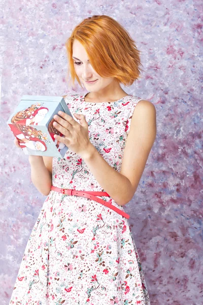 Vacker smal tjej i en kort klänning holding presentförpackning. Färgstark bakgrund. Porträtt av rödhåriga flicka — Stockfoto