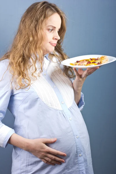 Krásná blondýnka těhotná žena s kouskem pizzy na talíři. Pizza na bílé plotně. Portrét těhotné ženy — Stock fotografie